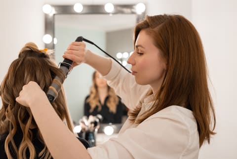 Side view of young female stylist using curling iron on hair of anonymous model near mirror in professional beauty salon