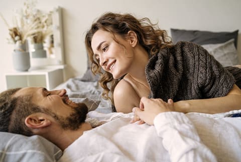 Delighted young man and woman smiling and looking at each other while resting on comfortable bed on weekend day at home