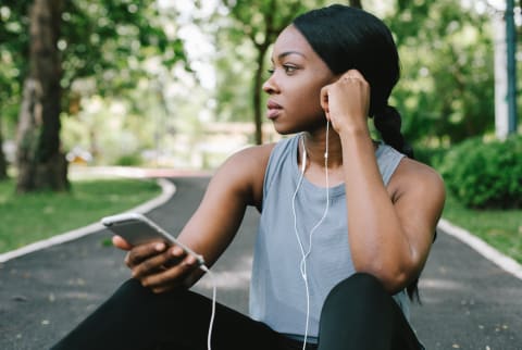A young black woman using her smart phone while listening to music and resting from a workout run in the park on the road amidst green grass and trees