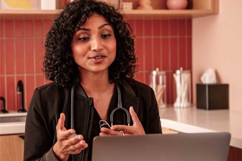 A female healthcare professional with dark, curly hair is engaged in a virtual consultation, speaking and gesturing with her hands while looking at a laptop screen. She is wearing a black jacket and has a stethoscope draped around her neck. The background is a warm-toned clinic space with a tiled, reddish-pink backsplash and wooden shelving with various objects.