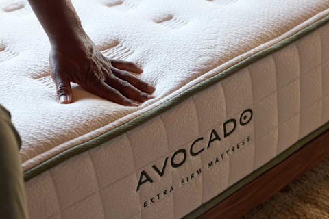 The image shows a person's hand gently pressing down on a quilted, light-colored mattress. The mattress has green piping along the edge, where the brand name "AVOCADO" and the product name "EXTRA FIRM MATTRESS" are clearly visible. The mattress is resting on a wooden bed frame.