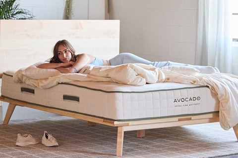 A woman is shown lying on her stomach, propped up on her elbows, on an Avocado Extra Firm Mattress. She is looking directly at the camera. A comforter and pillows are on the bed with her, and a pair of white sneakers are on the floor in front of the light wood bed frame. The brand name "AVOCADO" and the product description "EXTRA FIRM MATTRESS" are clearly visible on the side of the mattress.