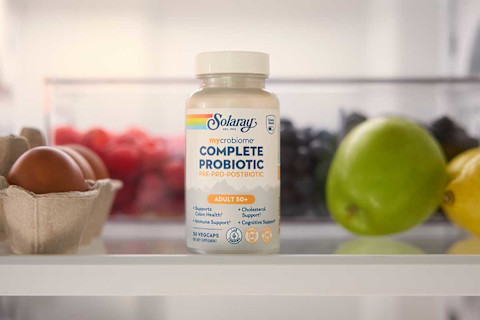 Alt Text: A bottle of Solaray mycrobiome Complete Probiotic for adults over 50 is on a shelf inside a refrigerator. The bottle is positioned in the middle of the shelf with fresh food, including a carton of eggs and various fruits, blurred in the background on either side.