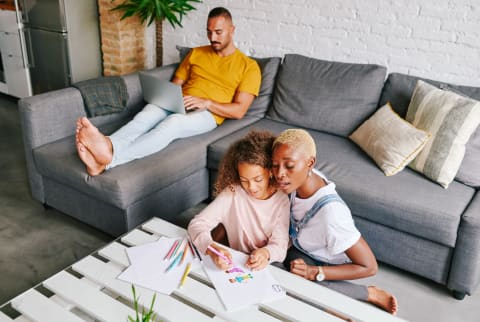 miling mother and her little daughter drawing pictures together at their coffee table with dad relaxing on the living room sofa