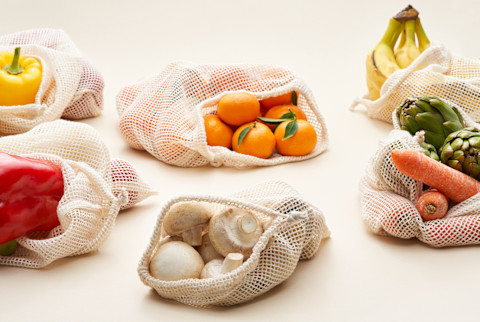 Assorted fresh fruits and vegetables in reusable cotton bags placed on cream background