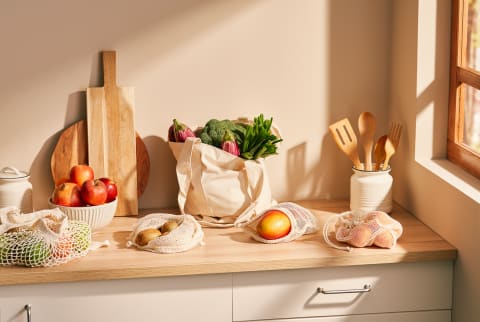 Bunch of various ripe fruits and vegetables in zero waste sacks placed on counter near wooden kitchenware against beige wall in contemporary kitchen