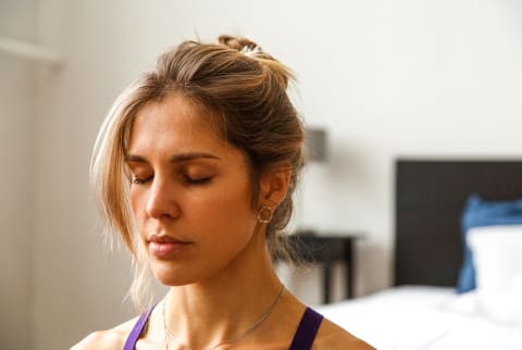 Calm young lady with closed eyes doing respiratory warm up exercises in bedroom at home