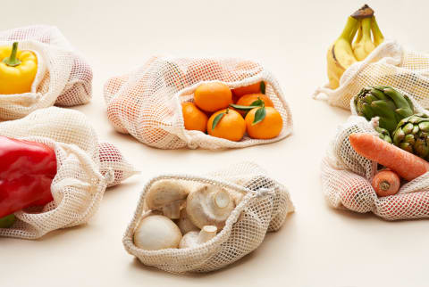 Assorted fresh fruits and vegetables in reusable cotton bags placed on cream background