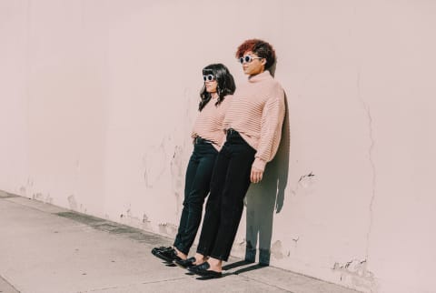 Two matching girls in pink sweaters, black jeans, and white sunglasses. These best friends are learning against a wall.