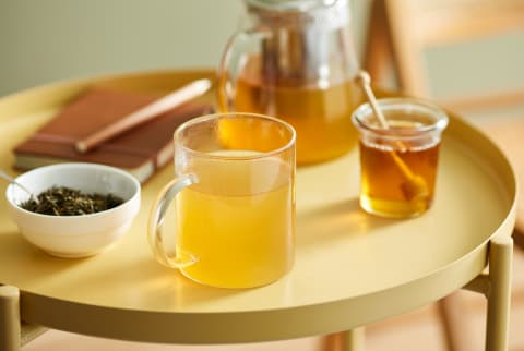 Close-up of sweet herbal tea with honey served in glass mug on round metallic table with bowl of dry leaves, notebook, jar of honey and glass teapot