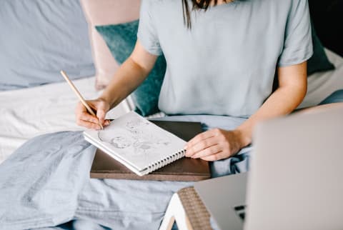 Unrecognizable female sitting on comfortable bed and drawing sketch in notepad while working at home