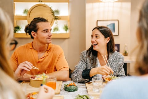 Man with bun and orange shirt sitting with brunette eating asian food.