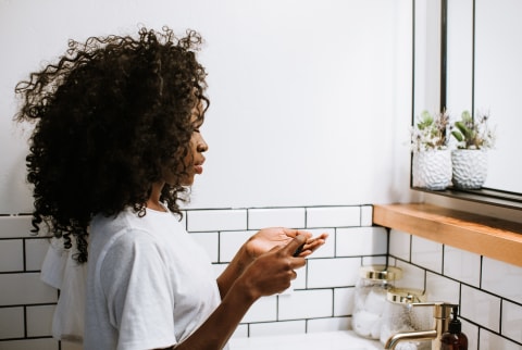 Woman Applying Skin Care Product In Bathroom Mirror
