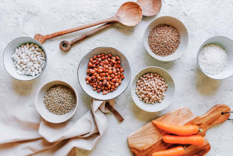 Flat lay of various beans & grains like kidney, lentils, chickpeas, white beans, farro, and white rice with wooden spoons and carrots