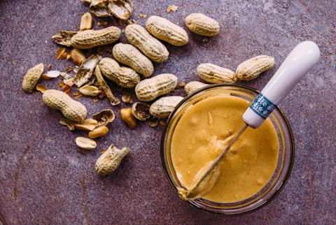 Overhead of peanut nut butter with raw peanuts on the side on top of purple background Overhead of peanut nut butter with raw peanuts on the side on top of purple background