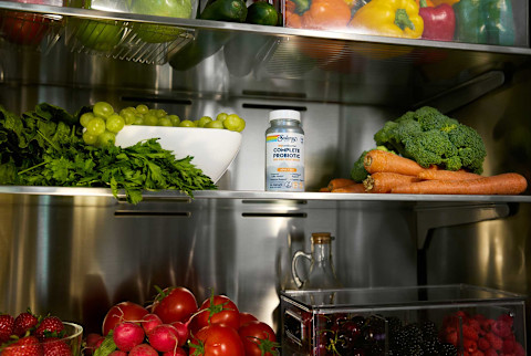 An open refrigerator is shown, with a bottle of Solaray mycrobiome Complete Probiotic for adults over 50 placed on a middle shelf. The shelves are fully stocked with a variety of fresh fruits and vegetables, including bell peppers, grapes, carrots, broccoli, and tomatoes.