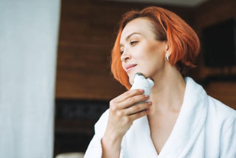 Beauty portrait of smiling woman 35 year in white bathrobe with clean fresh face and hands with red hair doing fasial massage with microcurrent facial massager at bath room, treat yourself, home body care