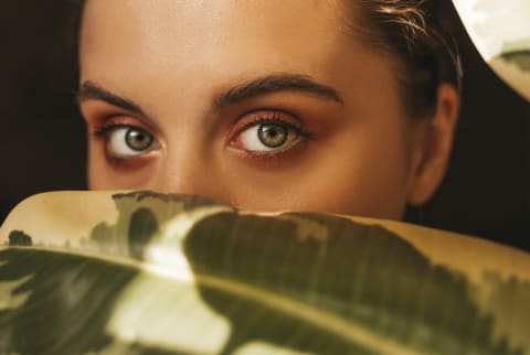 young woman with long eyelashes hiding behind a leaf