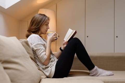 Woman drinking from a mug and reading a book on the couch.