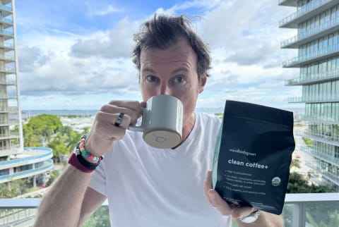 man outside on sunny day drinking coffee on balcony holding up bag of beans