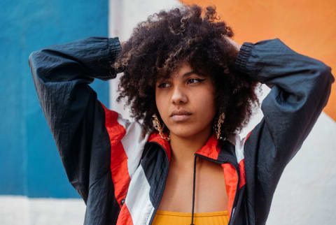 Woman in colorful jacket stands in front of a mural with her hands on her head and looks into the distance with worry on her face