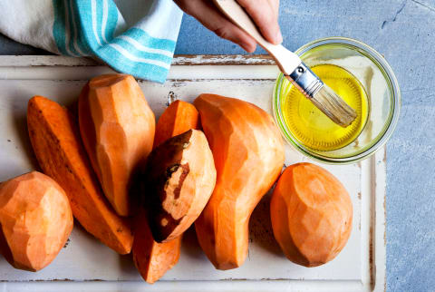 Overhead of peeled sweet potatoes with someone holding a brush over bowl of olive oil