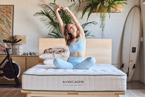 A woman in a light blue athletic outfit sits cross-legged on an Avocado Extra Firm Mattress with her arms stretched high above her head. The mattress, which has a quilted top and green piping, rests on a light wood bed frame. The surrounding room is a bright, loft-style space decorated with potted plants, framed art, a stationary bike, and a surfboard.