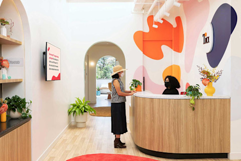 The reception area of a modern, brightly lit clinic. A woman in a sunhat and checkered shirt stands at a curved, wood-paneled reception desk speaking to an employee with a face mask. The wall behind the desk is decorated with large, abstract shapes in orange, pink, and navy blue, featuring the Tia logo. The room has light wood flooring, a red circular rug, arched doorways leading to other rooms, and shelves with plants and décor.