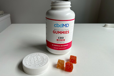 CbdMD Broad-spectrum gummies bottle open on white table next to stack of three gummies in red, orange, and yellow