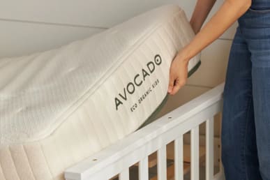 An Avocado Eco Organic Kids Mattress is fixed in the crib with a hand