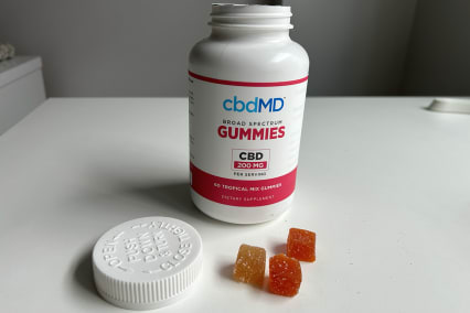 CbdMD Broad-spectrum gummies bottle open on white table next to stack of three gummies in red, orange, and yellow