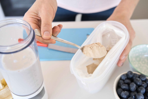 Womab scooping a powder supplement into a blender Womab scooping a powder supplement into a blender