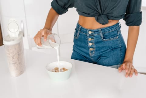 Anonymous female in casual outfit pouring fresh milk into bowl with cereal corn flakes while preparing healthy breakfast at home