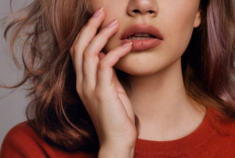 Partially obscured woman with full lips in a soft matte neutral lip color touches her fingers to her face.