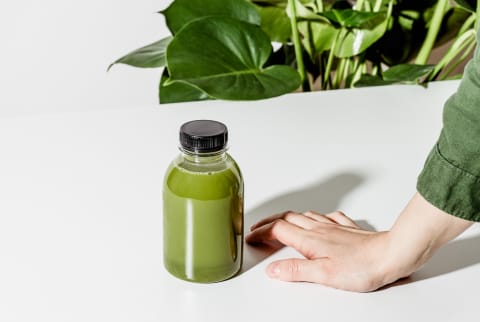 Hand Beside a Bottle of Green Juice on a White Table