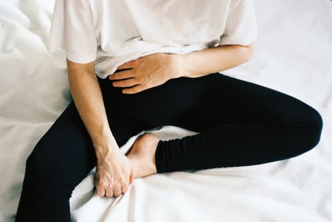 Young woman sitting on the white bed, holding her stomach and toes.