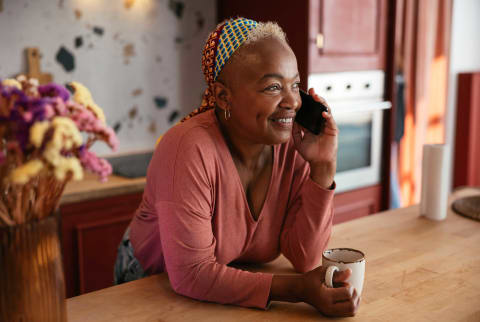 Mature woman talking on her cellphone in the kitchen drinking a mug of coffee