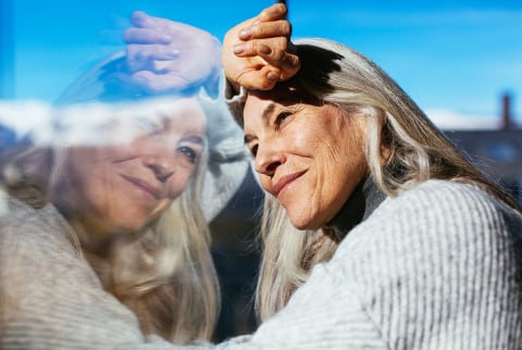 Cheerful mature lady with gray hair smiling and looking away while leaning on window and enjoying sunny day at home