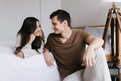 Couple posing together, brunette woman in bed smiling at her partner, male partner on the ground looking back at her.