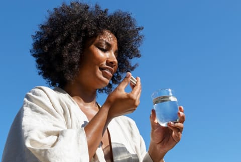 Black woman taking two multivitamin capsule holding a glass of water with a blue sky in the background