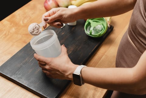 Athletic woman in sportswear with measuring spoon in her hand puts portion of whey protein powder into a shaker on wooden table with amino acid white capsules, bananas and apple, making protein drink.
