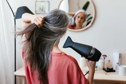 Mature woman blowdries grey hair in front of mirror