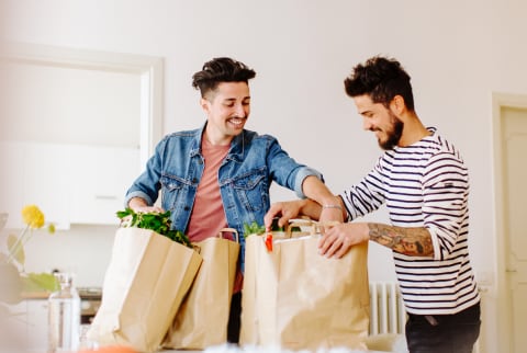 A couple smiling and unpacking paper bags with food in cozy room in contemporary apartment