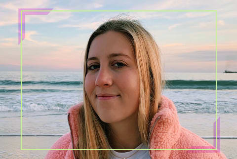 Blonde girl wearing a pink fuzzy jacket and a white tshirt, standing in front of an ocean