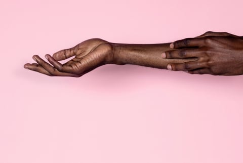 Black person running their fingertips against their outstretched arm against a pink background. 