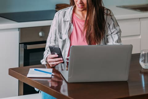 Brunette woman wearing pink shirt with sitting in front of a computer looking at her phone