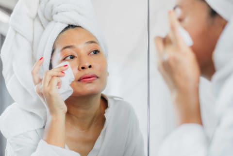 Asian woman with hair in a towel looks at skin up close in mirror