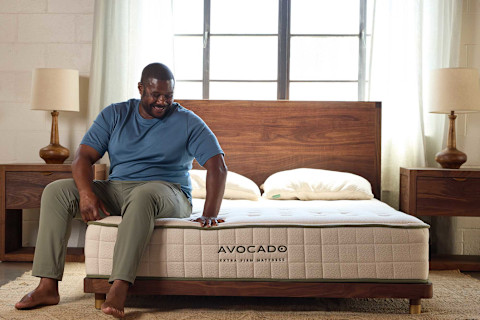 A smiling man sits barefoot on the edge of an Avocado Extra Firm Mattress, resting his hand on the quilted surface. The mattress, which has green trim, is set on a dark wood bed frame with matching wood nightstands and lamps on either side.