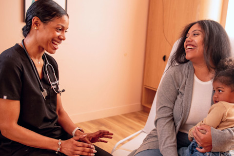 A healthcare provider in black scrubs and a stethoscope sits smiling, facing a laughing female patient who is holding a young child on her lap. All three subjects are engaged in a positive conversation within a clinic or exam room setting with pale pink walls.