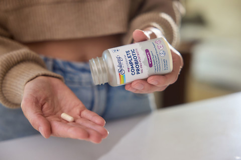  The image shows a person's hands holding a bottle of Solaray mycrobiome Complete Probiotic for Women's health. The bottle's label highlights support for "Vaginal Health," "Urinary Health," and "Daily & Occasional Stress Support." In the palm of the person's other hand, a single, light-colored capsule is shown. The person is wearing a beige knit sweater and is standing over a white surface.